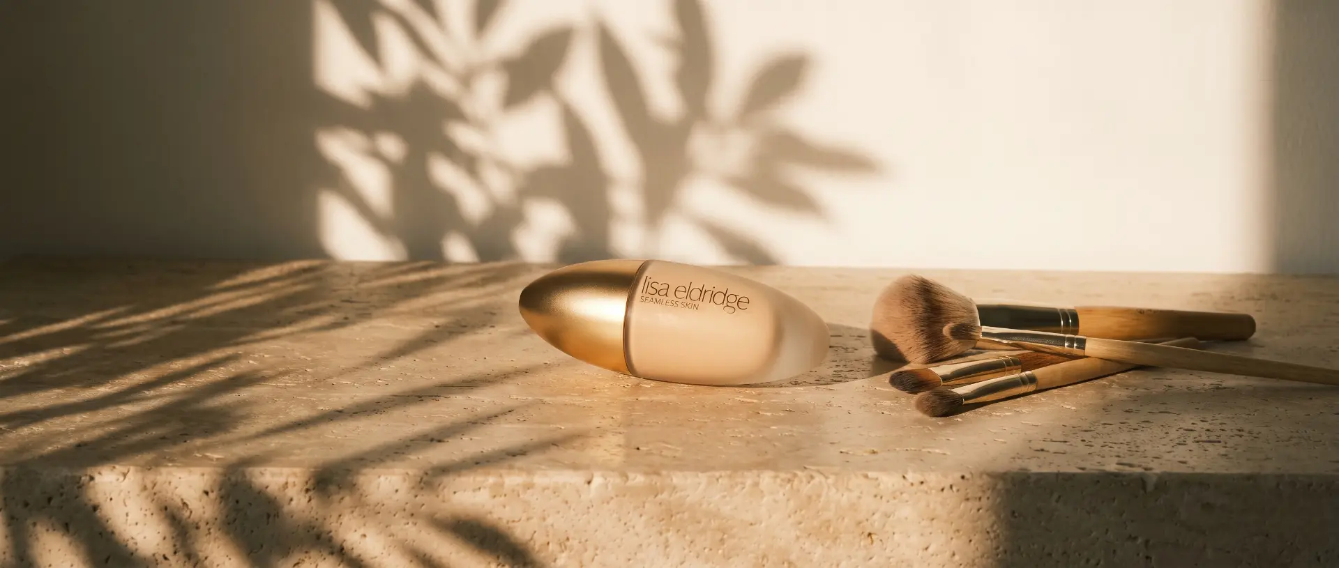 Lisa Eldridge Seamless Skin Foundation in goldener Glasflasche neben Make-up-Pinseln auf warmem Naturstein im Sonnenlicht