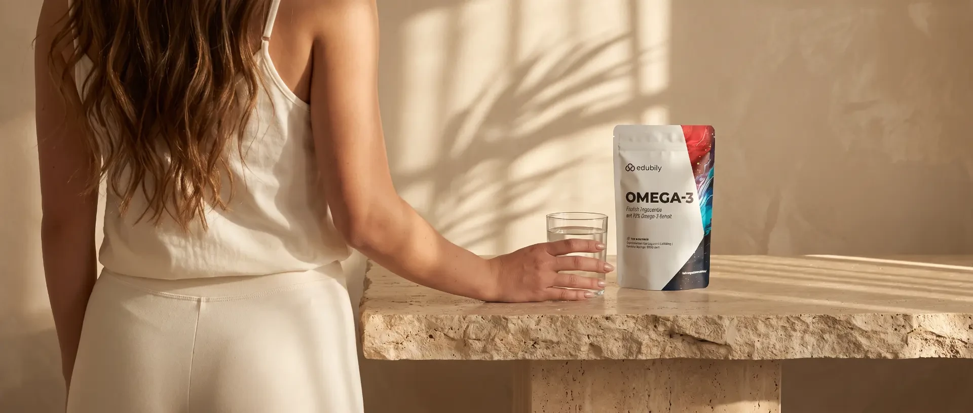 Eine Frau mit einem Wasserglas neben einer Packung edubily Omega-3 Kapseln auf einer Natursteinfläche